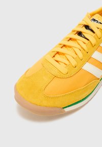 Gul sneaker med ovandel i mocka och textil, med vita ränder och en grön accent längs sulan. Texturerat snörningsområde.