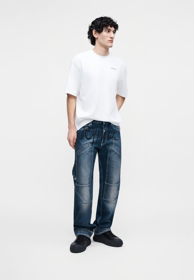 Jeune homme aux cheveux bouclés, portant un t-shirt blanc oversize, un jean bleu marine ample avec de grandes poches, et des baskets noires.