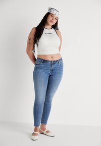White cropped tank top with black lettering, blue skinny jeans, white flat shoes with polka dots, and a patterned headband.