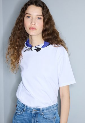 Junge Frau mit langen lockigen Haaren, die ein weißes T-Shirt, blaue Jeans und ein blau-schwarz gemustertes Halstuch trägt, steht vor einem grauen Hintergrund.