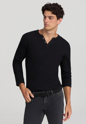 Maglietta henley nera a maniche lunghe realizzata in tessuto a coste, dotata di una placca a quattro bottoni e di un design aderente, abbinata a jeans scuri e a una cintura intrecciata.