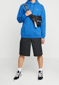 Blå hoodie, svarta shorts och svarta sneakers. Klädseln inkluderar en svart crossbody-väska med ett vitt logoband och silverdetaljer. 