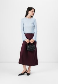 Žena stojí čelem vlevo, má na sobě světle modrý žebrovaný top a vínovou sukni, drží malou černou kabelku.