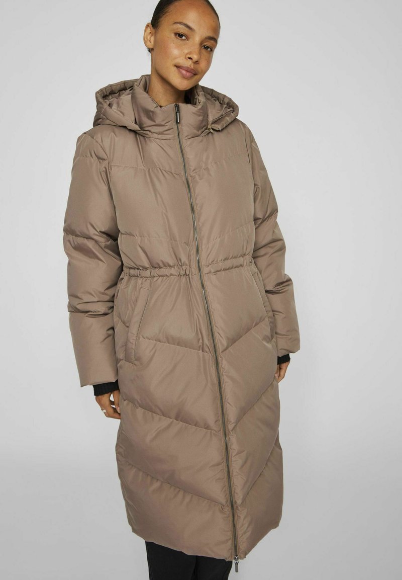 Manteau long rembourré de couleur marron clair avec capuche, fermeture éclair à l'avant, taille élastique et motif piqué diagonal. Texture douce et lisse avec poches latérales.