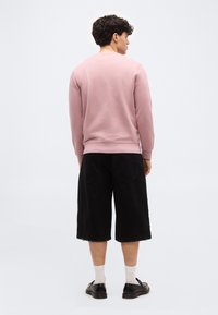 Άντρας γυρισμένος με την πλάτη φοράει ροζ φούτερ, μαύρα φαρδιά σορτς έως το γόνατο, λευκές κάλτσες και μαύρα παπούτσια πάνω σε ένα απλό ανοιχτό φόντο.