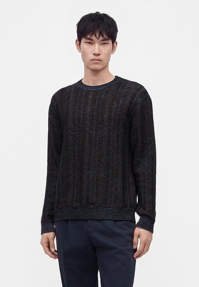 Jeune homme aux cheveux courts noirs portant un pull noir texturé à manches longues et un pantalon foncé sur un fond uni clair.
