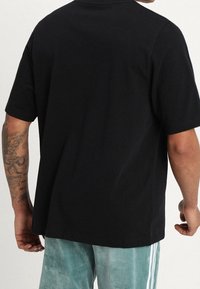 Černé bavlněné tričko s krátkými rukávy, kulatým výstřihem a rovný lem. Viditelné švy; nošeno s světlými kalhotami.