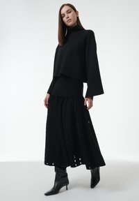 Vrouw in een zwarte coltrui, textuur midi-rok en zwarte geklede enkellaarsjes, staande tegen een effen witte achtergrond.