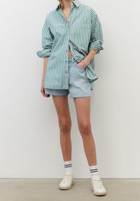 Camicia over size a righe verdi e bianche, pantaloni corti in denim, sneakers bianche con righe nere sui calzini. Outfit informale con una vestibilità rilassata.