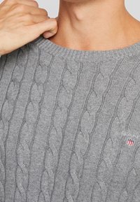 Szary sweter z dzianiny z prążkowanym okrągłym dekoltem, z wzorem w warkocze i małą naszywką z logo z czerwonymi i niebieskimi akcentami w okolicy kołnierza.