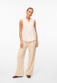 Top senza maniche color crema con scollatura a V e dettagli con bottoni, abbinato a pantaloni beige larghi, realizzati in un tessuto morbido con taglio dritto.