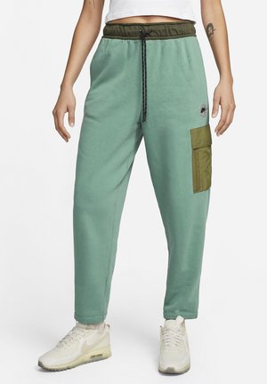 Personne portant un pantalon jogger cargo vert avec une poche olive et un cordon noir, des baskets blanches, et un top blanc court.