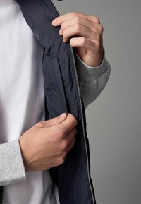 Veste marine avec une fermeture éclair métallique, montrée en train d'être dézippée. Le tissu intérieur est lisse, contrastant avec le pull gris au niveau du poignet.