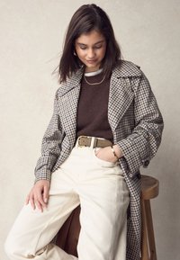 Plaid trench coat with wide collar, brown knit sweater, cream trousers, and textured belt. Gold jewelry accents complete the look.