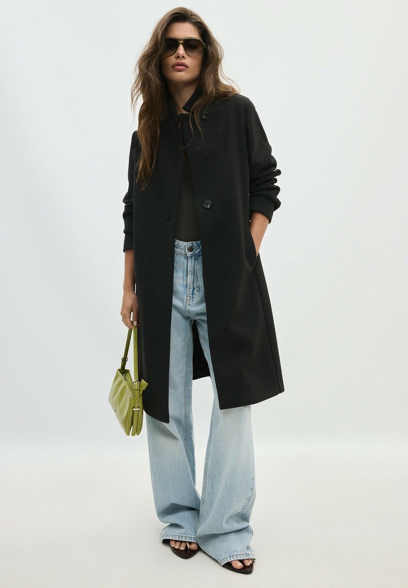 Femme aux longs cheveux bruns portant des lunettes de soleil foncées, un manteau noir, un jean large clair, des sandales, et tenant un petit sac à main vert.