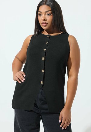 Vrouw met steil zwart haar draagt een mouwloos zwarte top met gouden knopen en een zwarte broek, poseert met hand op heup tegen een witte achtergrond.