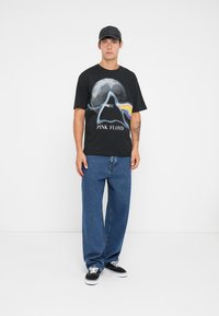 Svart bomull t-skjorte med grafisk design av et prisme og teksten "PINK FLOYD," kombinert med blå jeans og svarte sneakers.