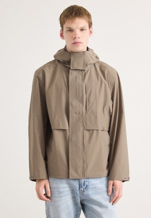 Veste imperméable marron clair avec un col haut, fermeture à pression à l'avant et deux grandes poches. Conçue pour un ajustement décontracté, texture minimale.