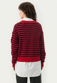 Chandail rayé rouge et marine avec un col rond, des poignets et un ourlet côtelés. Superposé sur une chemise blanche. Tissu en tricot texturé. Coucou décontracté.