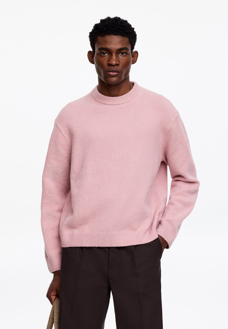 Jeune homme portant un pull en maille rose clair et un pantalon foncé, debout avec une main dans la poche et tenant un sac beige.
