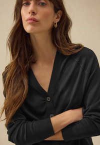 Cardigan noir avec un décolleté en V profond et des fermetures à boutons, présentant des manches longues et des poignets côtelés. Tissu doux avec une texture lisse.