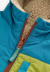 Chaqueta con capucha en azul y verde con una cremallera marrón, que presenta un forro suave en color beige y un tirador de cremallera decorativo con un patrón de chevron.