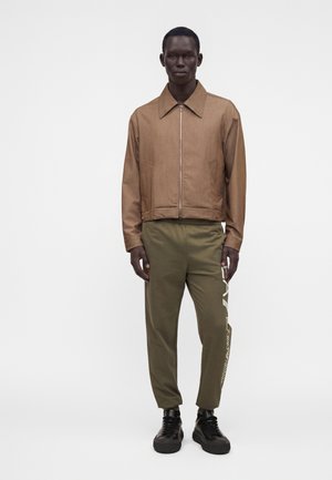 Hombre de pie con chaqueta marrón con cremallera, pantalones verde oliva con texto lateral y zapatillas negras sobre un fondo claro y liso.