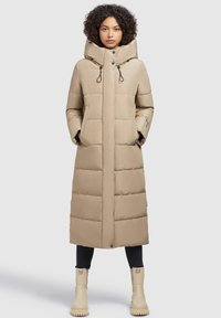 Sieviete, ģērbusies garā bēšā pildītā mētelī ar augstu apkakli, melnās biksēs un bēšos masīvos zābakos, stāv pretī vienkāršai gaišai fonam.