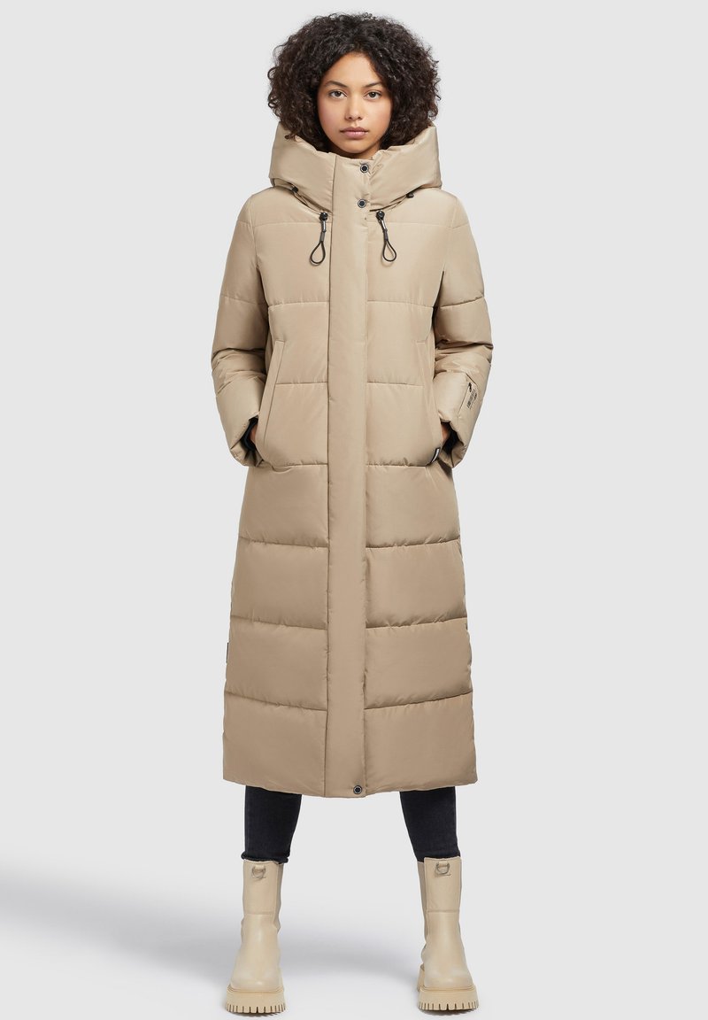 Sieviete, ģērbusies garā bēšā pildītā mētelī ar augstu apkakli, melnās biksēs un bēšos masīvos zābakos, stāv pretī vienkāršai gaišai fonam.