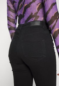 Jeans neri con un design aderente, dotati di un alto punto vita, tasche posteriori e cuciture visibili. Il materiale sembra elastico e liscio.