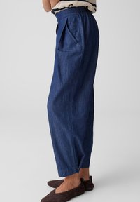 Woman wearing high-waist, loose-fitting dark blue denim pants with side pockets and brown slip-on shoes against a plain background.