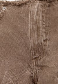 Lähikuva kuluneesta ruskeasta denimpäällyksestä, jossa näkyvät saumat ja metalliruuvi lähellä yläreunaa.