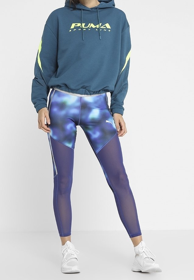 Tealfärgad hoodie med gula detaljer och grafisk logotyp. Lila leggings med blå abstrakt mönster och nätpaneler på sidorna. Vita sneakers.