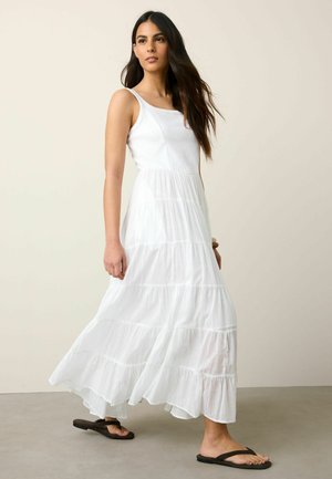 Frau, die in einem ärmellosen, stufigen weißen Maxikleid und schwarzen Flip-Flops vor einem einfarbigen beigefarbenen Hintergrund geht.