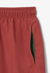 Pantaloni corti rossi in tessuto con vita elasticizzata, dotati di una tasca laterale foderata in verde. Texture liscia, design semplice con un colore uniforme.