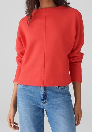 Femme portant un pull rouge à manches longues avec une couture centrale, associé à un jean denim bleu clair, sur un fond gris uni.
