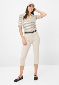 Beige gestreepte polo met V-hals, beige cropped broek, zwarte riem en zwarte mesh platte schoenen. Model draagt een beige pet.