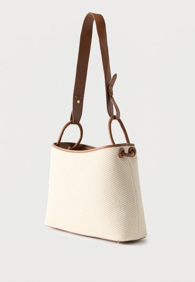 Μπεζ πλεκτή τσάντα χειρός με καφέ δερμάτινες λεπτομέρειες και ρυθμιζόμενο λουρί ώμου, απεικονισμένη πάνω σε απλό λευκό φόντο.
