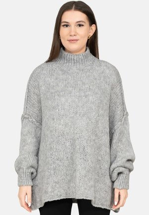 Kobieta ubrana w luźny, jasnoszary, grubo pleciony sweter z wysokim kołnierzem i długimi rękawami, stojąca na jednolitym tle.