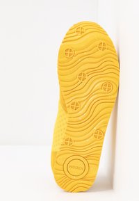 Gelbe Sneaker-Sohle mit einem wellenförmigen Muster, die kreisförmige Griffdetails und strukturierte Seiten aufweist. In der Mitte mit "WODEN" gekennzeichnet.