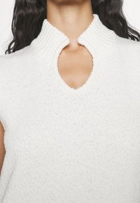 Top in maglia bianco senza maniche con colletto alto a costine e un taglio a chiave rotondo sul collo, realizzato in un tessuto testurizzato.