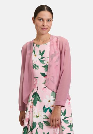 Giacca leggera e trasparente di colore rosa, con apertura frontale, maniche a sbuffo e design fluente. Indossata sopra un vestito a fiori in toni pastello.
