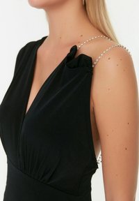 Robe noire avec un décolleté en V profond, présentant un tissu froncé sur le corps et des bretelles ornées de rhinestones scintillants. Texture lisse, design ajusté.