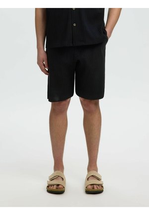 Osoba u crnim rebrastim kratkim hlačama i odgovarajućoj košulji, bež sandalama otvorenih prstiju, stoji s jednom rukom u džepu na bijeloj pozadini.