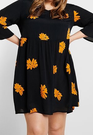 Sort løstsiddende kjole med orange blomsterprint, trekvartede ærmer, båret af en person med hænderne på hofterne mod en ensfarvet baggrund.