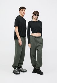 Fekete kerek nyakú póló és hosszú ujjú felső laza, olívazöld színű szabadidőnadrággal párosítva. Mindkét modell vastag talpú fekete tornacipőt visel.