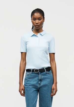 Vrouw staand, gekleed in een lichtblauw poloshirt met korte mouwen, ingestopt in een high-waisted blauwe spijkerbroek met een zwarte riem, tegen een effen achtergrond.