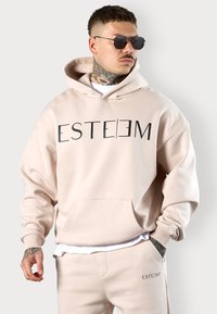 Helle rosa Kapuzenjacke mit dem Wort "ESTEEM" in fetten schwarzen Buchstaben. Verfügt über eine Fronttasche und eine entspannte Passform. Das Modell trägt zudem passende Hosen.