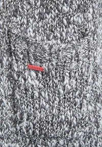 Detailní záb ěr na šedou a bílou pletenou tkaninu s malou červenou visačkou s nápisem "GC&L", přišitou blízko texturovaného švu.