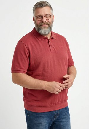 Lächelnder bärtiger Mann mit Brille, der ein rotes Poloshirt und blaue Jeans trägt, steht mit verschränkten Händen vor einem schlichten weißen Hintergrund.